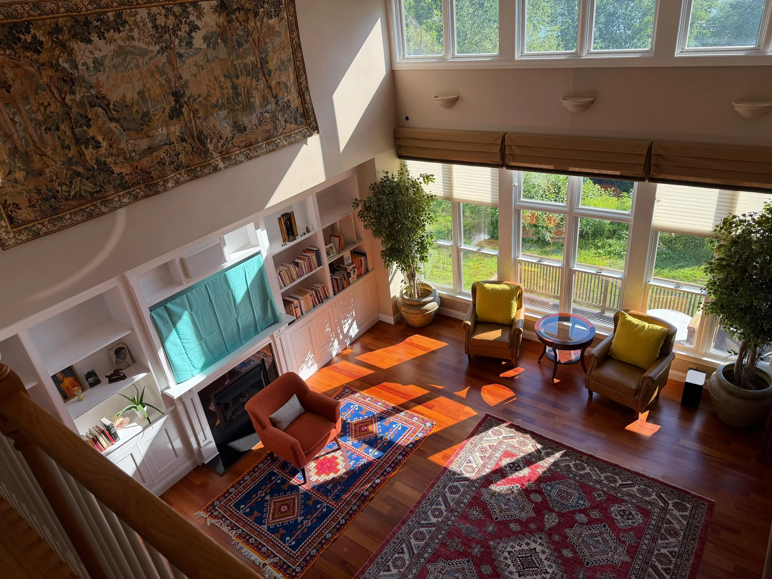 A bright living room with large windows, wooden floors, a Persian-style rug, bookshelves, and seating with yellow and orange cushions.