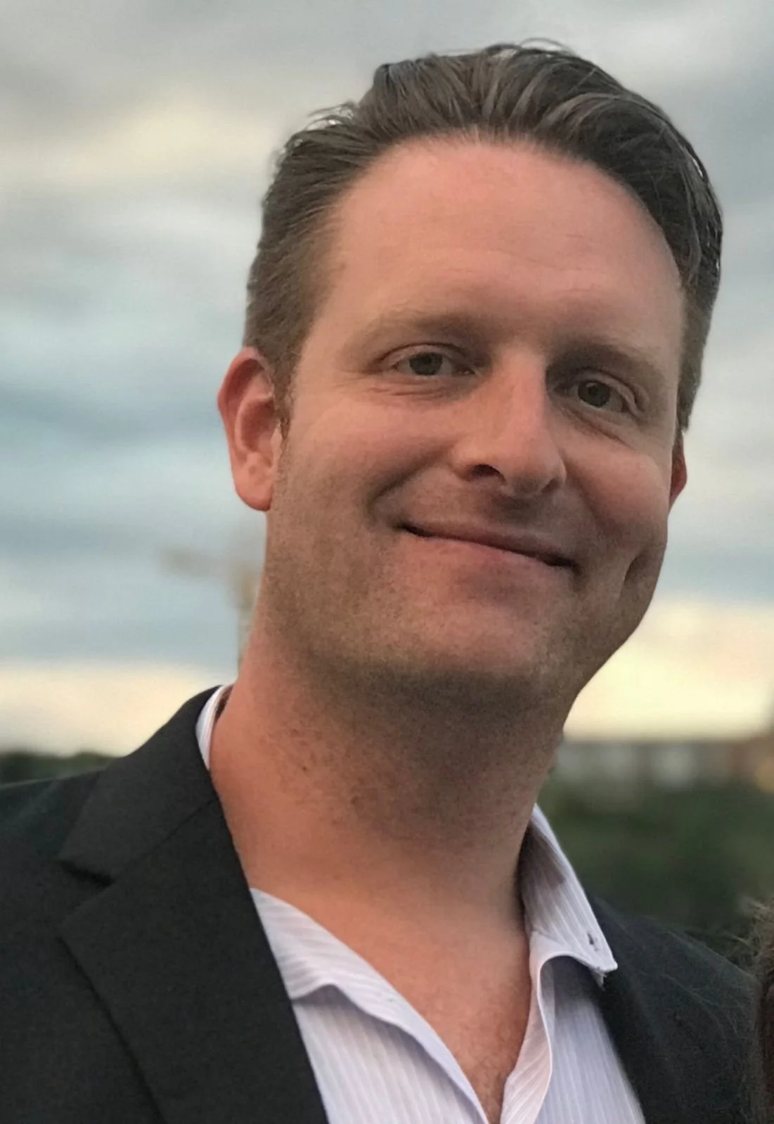 A smiling man in a suit jacket with a cloudy sky background.