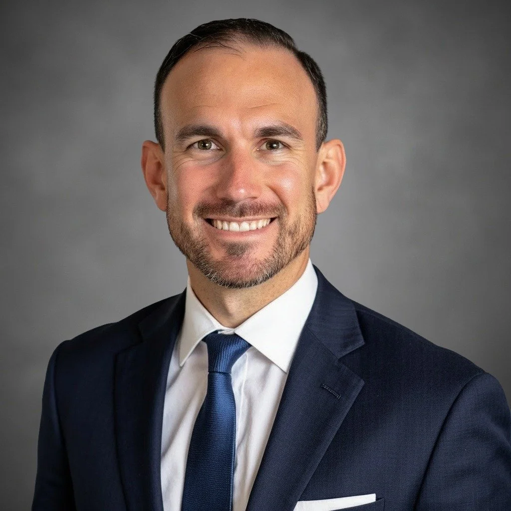 professional headshot of Gaston Escudero III wearing professional navy blue blazer and blue tie on gray background