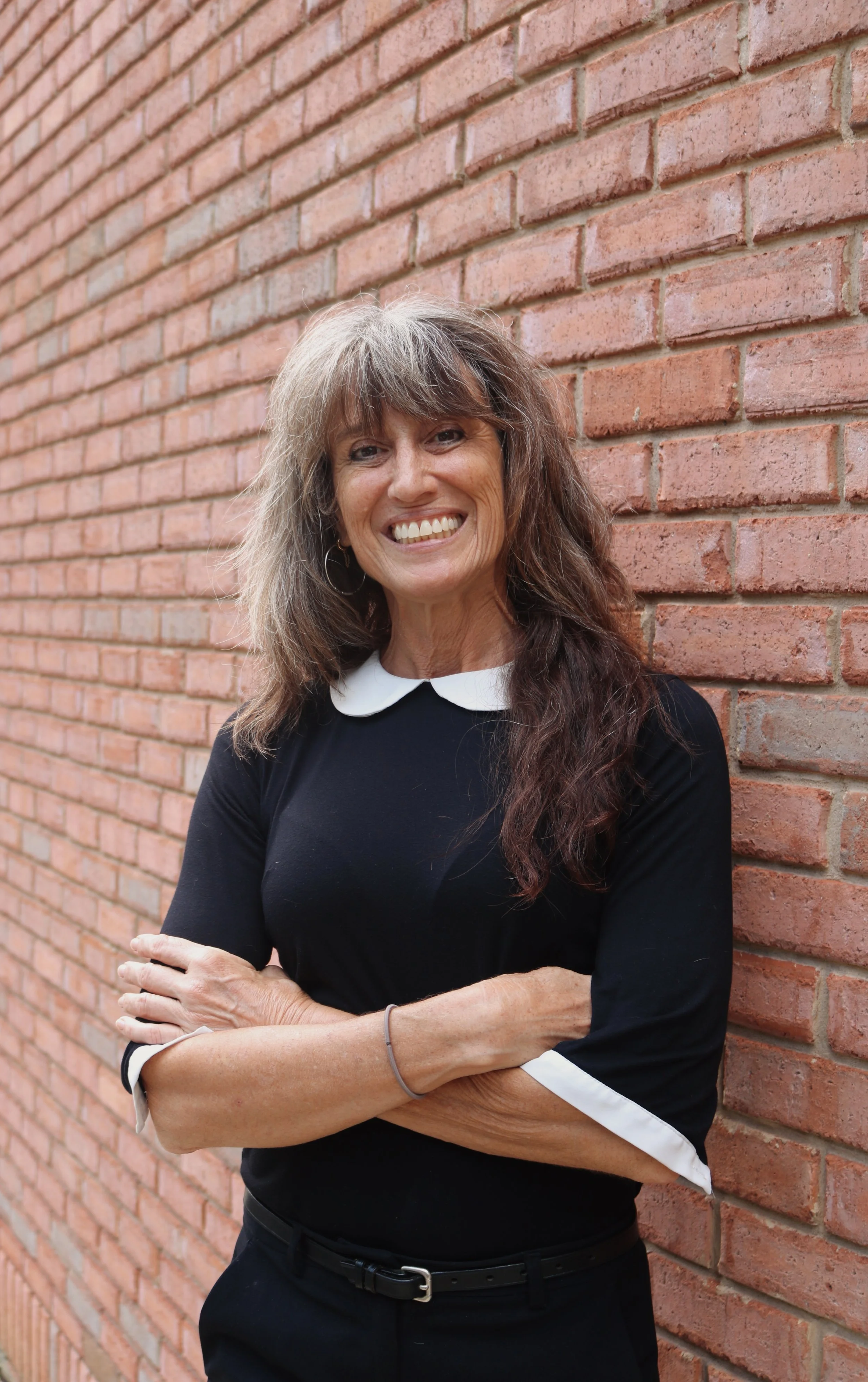 Great looking middle-aged woman with gray and brown hair smiling, standing against a red brick wall with her arms crossed, wearing a black top with white cuffs looking sharp, intelligent and ready to have your attention.