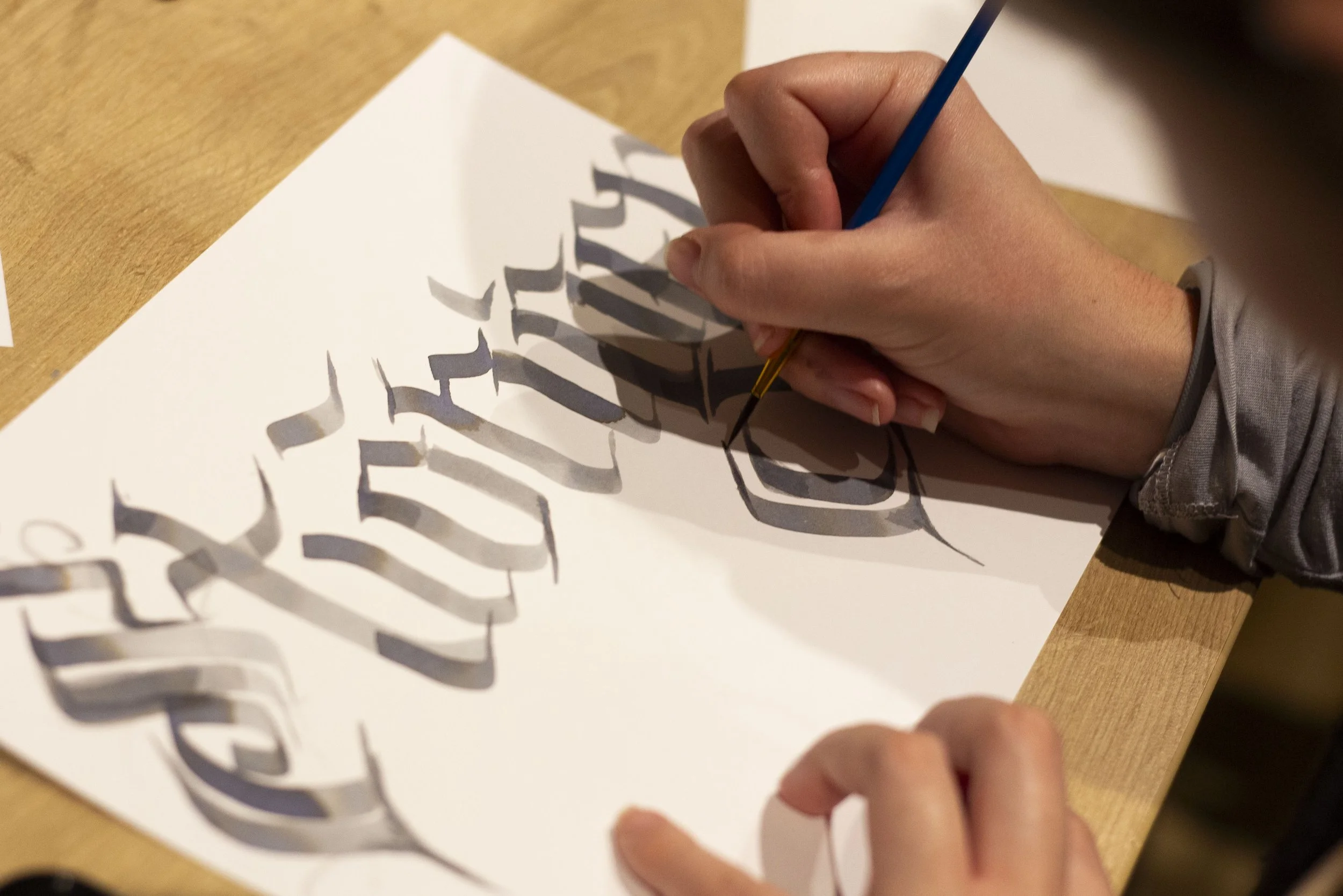 A person using a small paintbrush to paint black calligraphy on a white paper with a pattern of cut-out paper strips creating shadow effects.