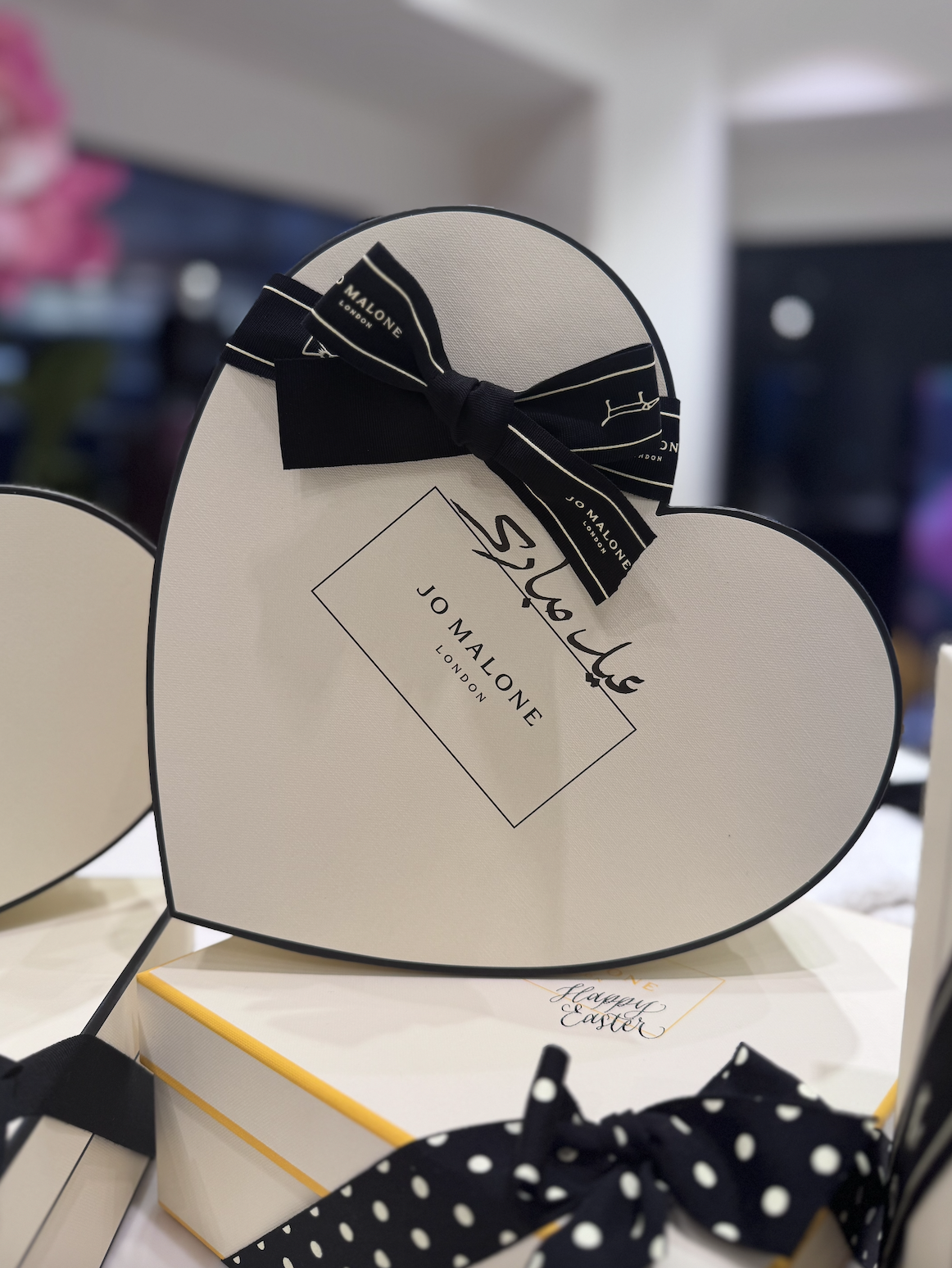 A white heart-shaped gift box with a black ribbon featuring the Jo Malone London logo, displayed in a store setting, with some other gift boxes visible in the background.