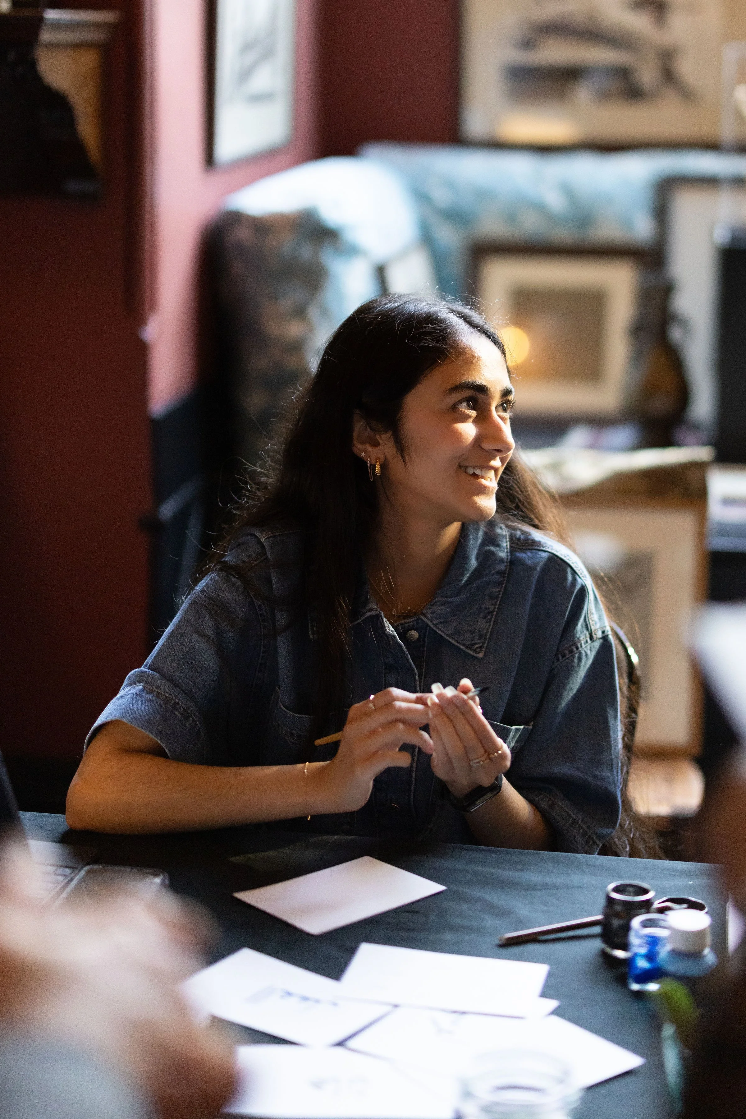 A young woman with long dark hair, wearing a denim jacket, sitting at a table with art supplies, smiling and looking to her left in a cozy indoor setting with artwork on the walls.