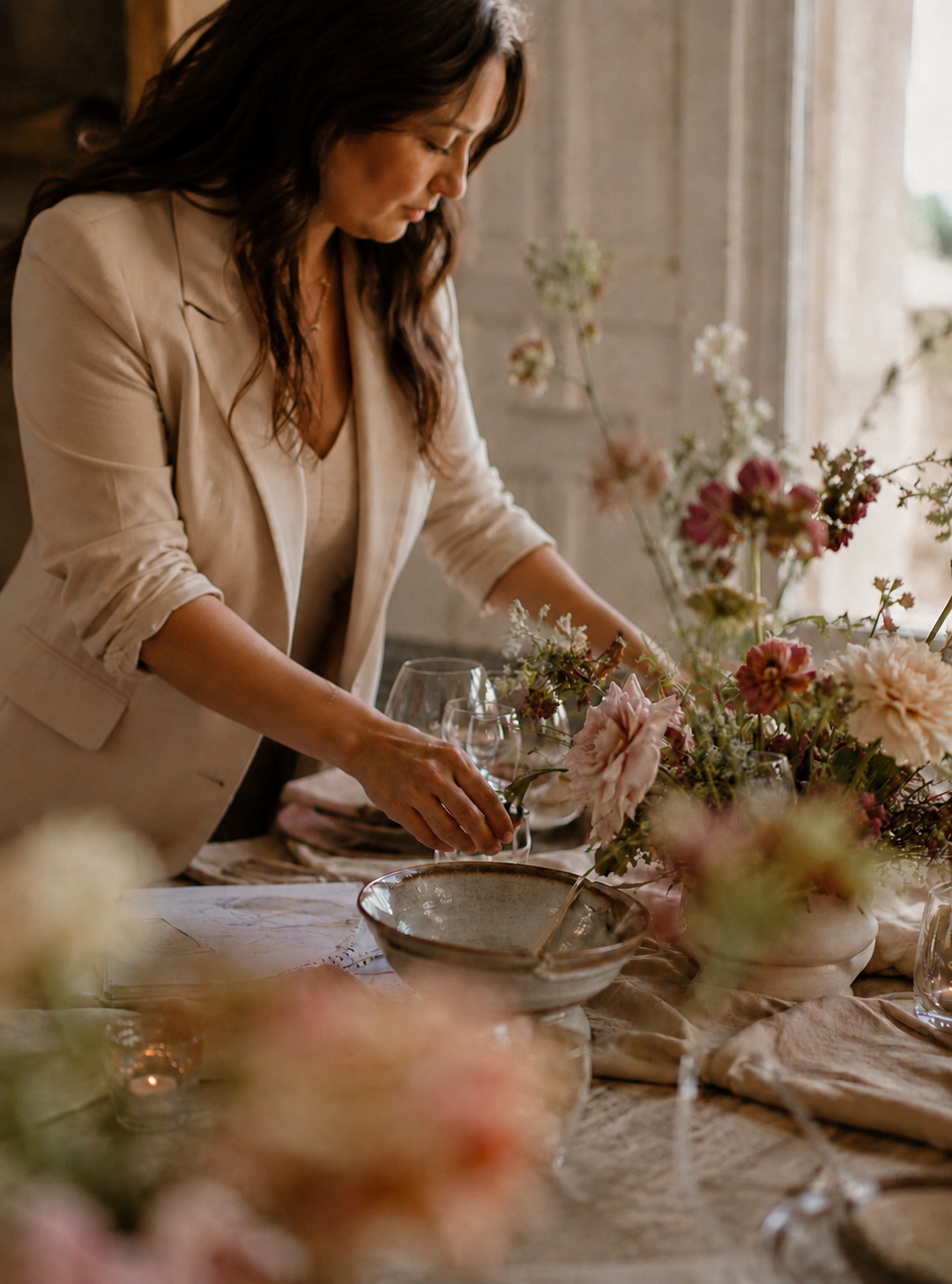 A woman arranging a floral centerpiece on a dining table with elegant glassware and a large floral arrangement in a softly lit room.