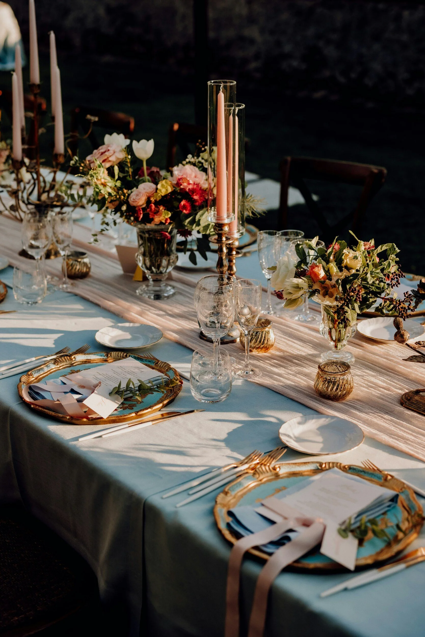 A elegantly set outdoor dining table decorated with floral arrangements, candles, and fine tableware for a formal event or celebration.