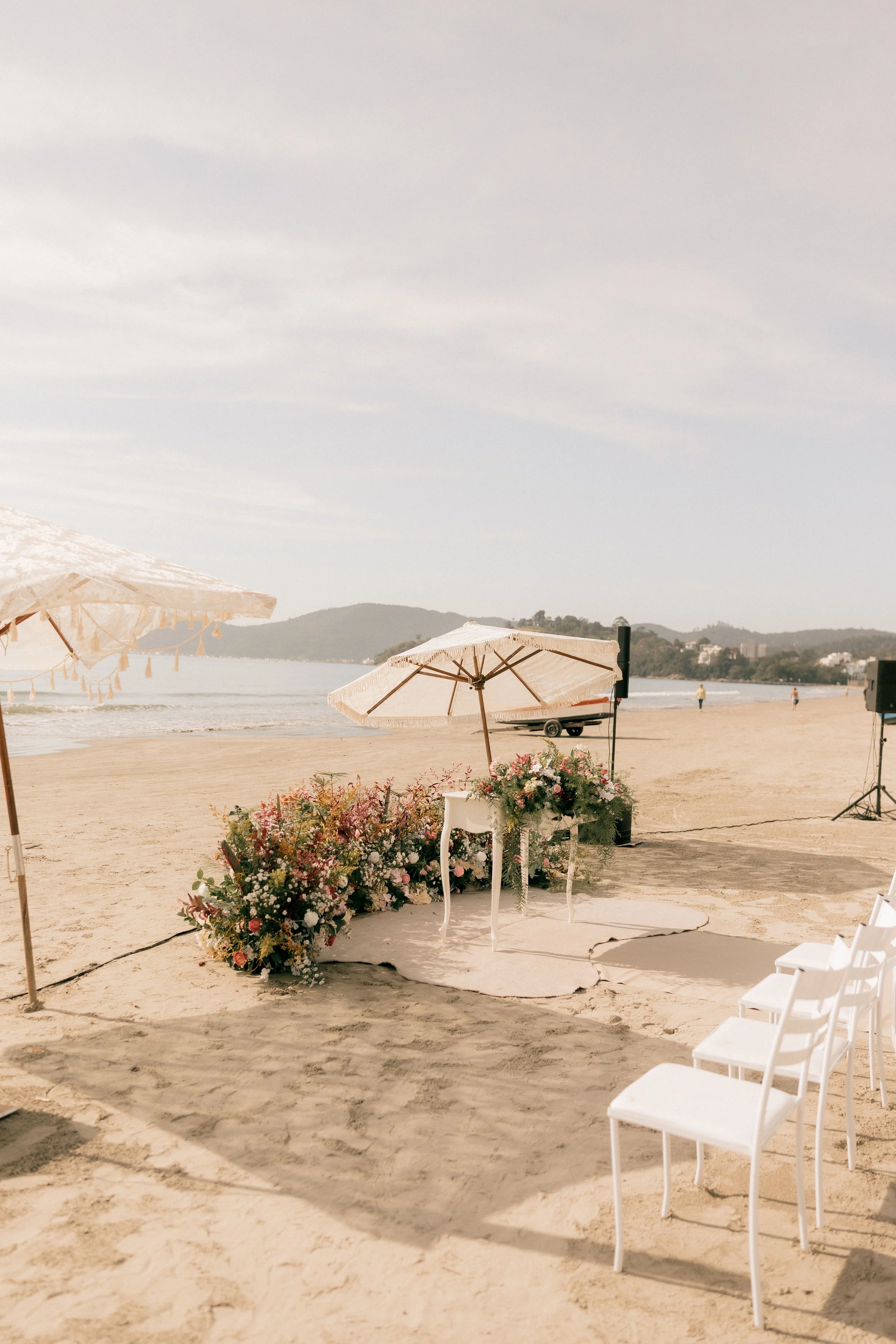 Beach setup with white chairs and floral arrangements in front of an umbrella, overlooking the ocean and distant hills under a cloudy sky.