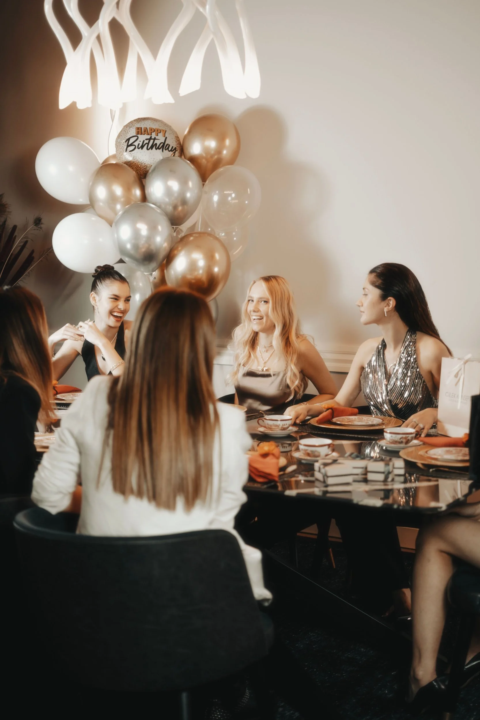 A birthday celebration with women sitting around a table, smiling and enjoying each other's company. There is a cluster of balloons in metallic and white colors with a 'Happy Birthday' balloon on top, and a modern white chandeliers above the group.