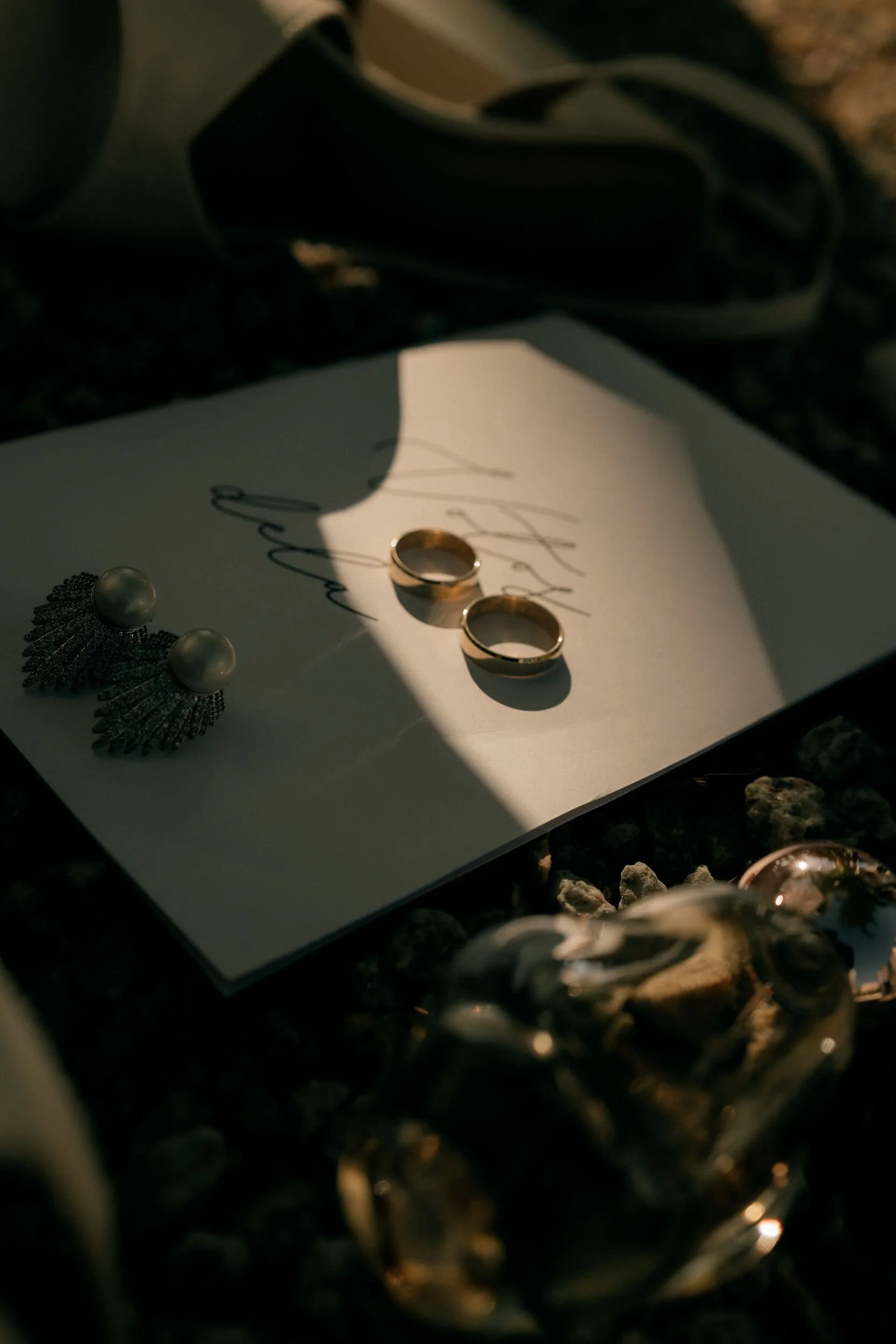 A pair of gold wedding rings resting on a white card with handwritten text, a pair of pearl earrings with black feather-like details, a black box or case, and some rocks or gravel around it, with a shadow cast over part of the scene.