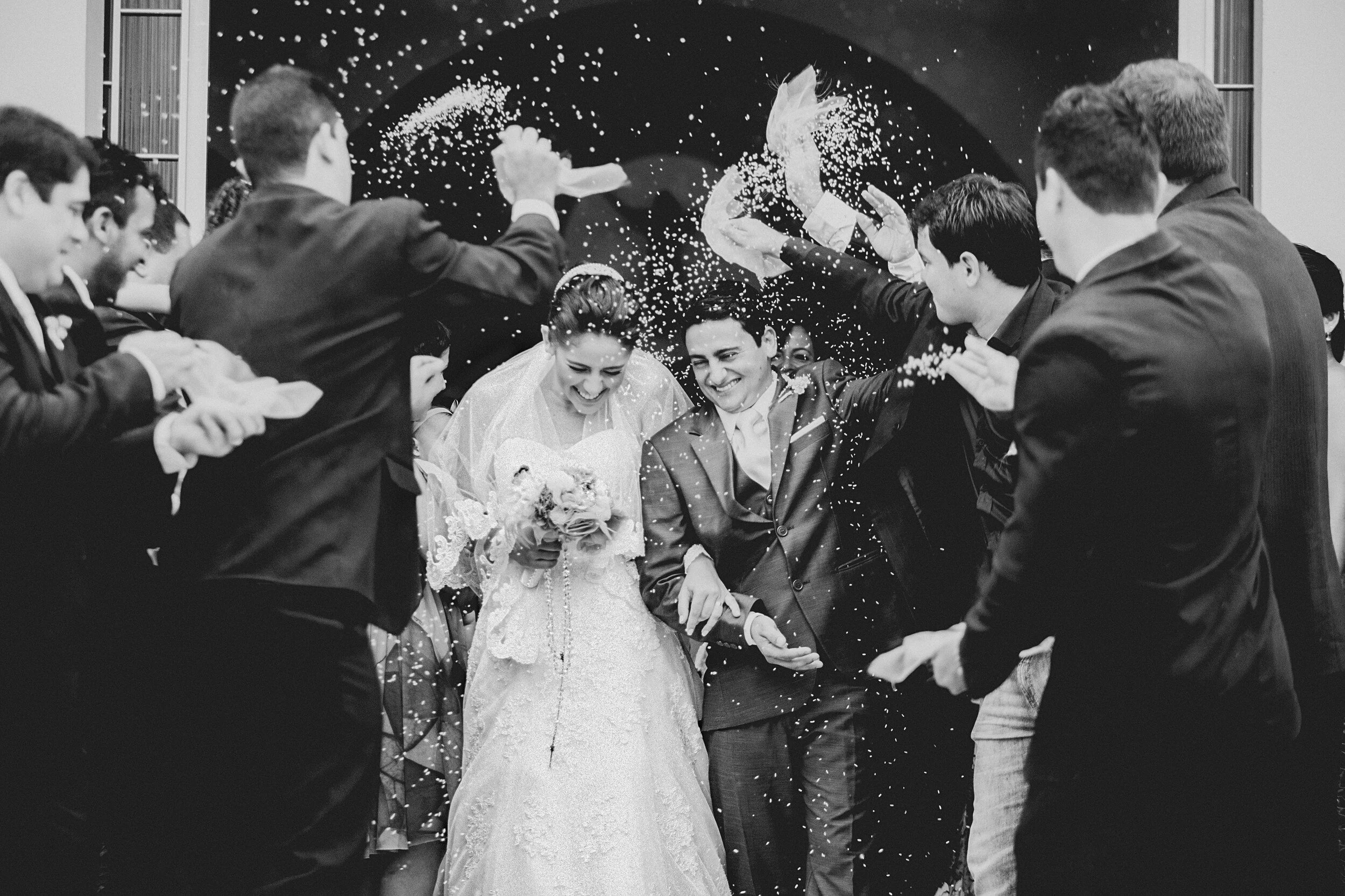 Black and white photo of a bride and groom smiling and celebrating with guests tossing confetti in the air during a wedding reception.
