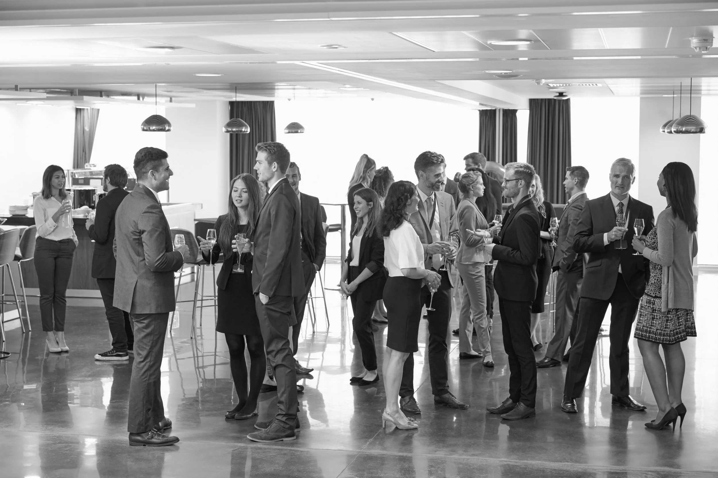 Group of well-dressed men and women socializing with drinks in a modern, spacious lounge or reception area, some engaged in conversations, with high tables, chairs, and large windows with blinds.