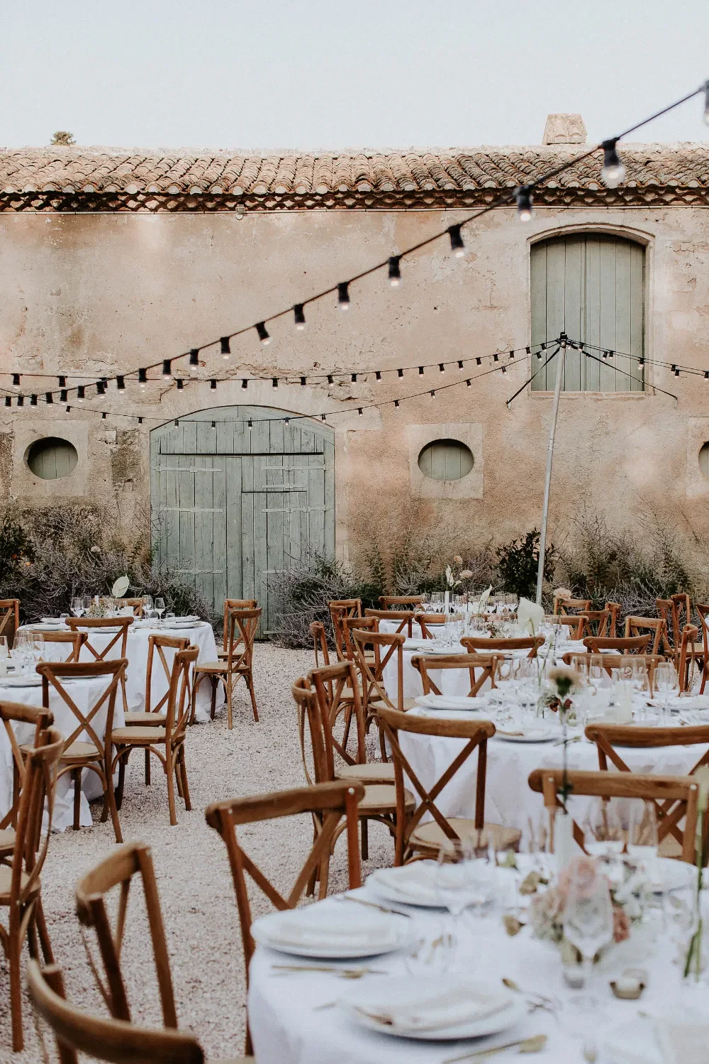 Outdoor dining setup with round tables and wooden chairs, decorated with white tablecloths, flowers, and tableware, in front of an old stucco building with closed green doors and string lights overhead.