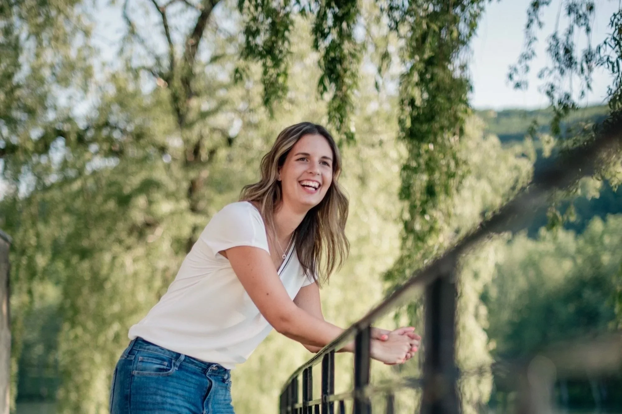 A woman with long brown hair, wearing a white T-shirt and blue jeans, leaning on a black railing outdoors in a park with green trees and hills in the background, smiling and looking off to the side.