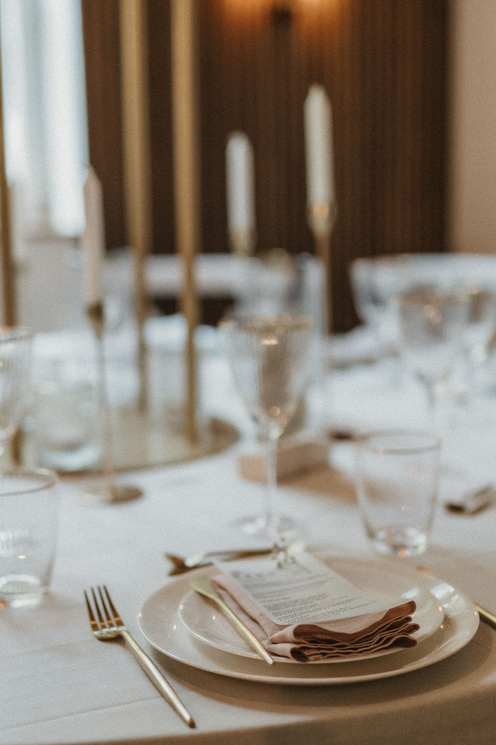 A formal dining table set with a white plate, gold fork and knife, beige napkin, and a menu card, with elegant glassware and a candelabra in the background.