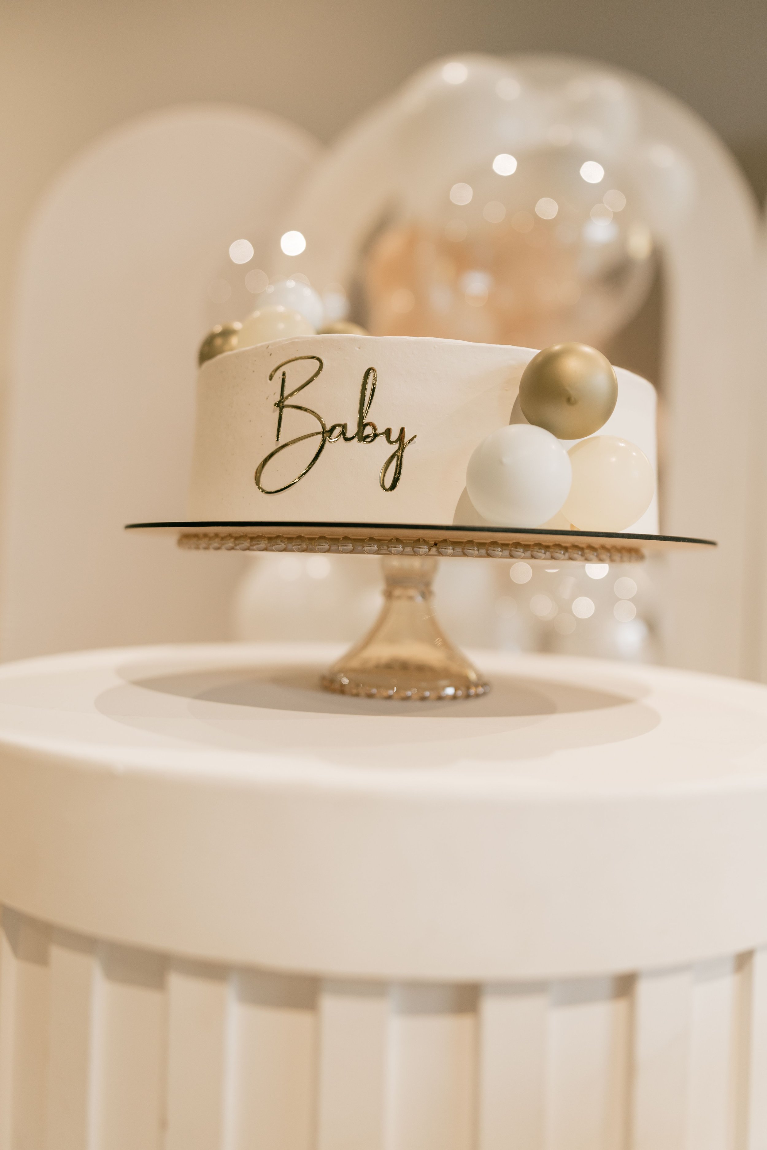 A white cake with gold and white spherical decorations and the word "Baby" written on it, displayed on a white cake stand with a decorative base.