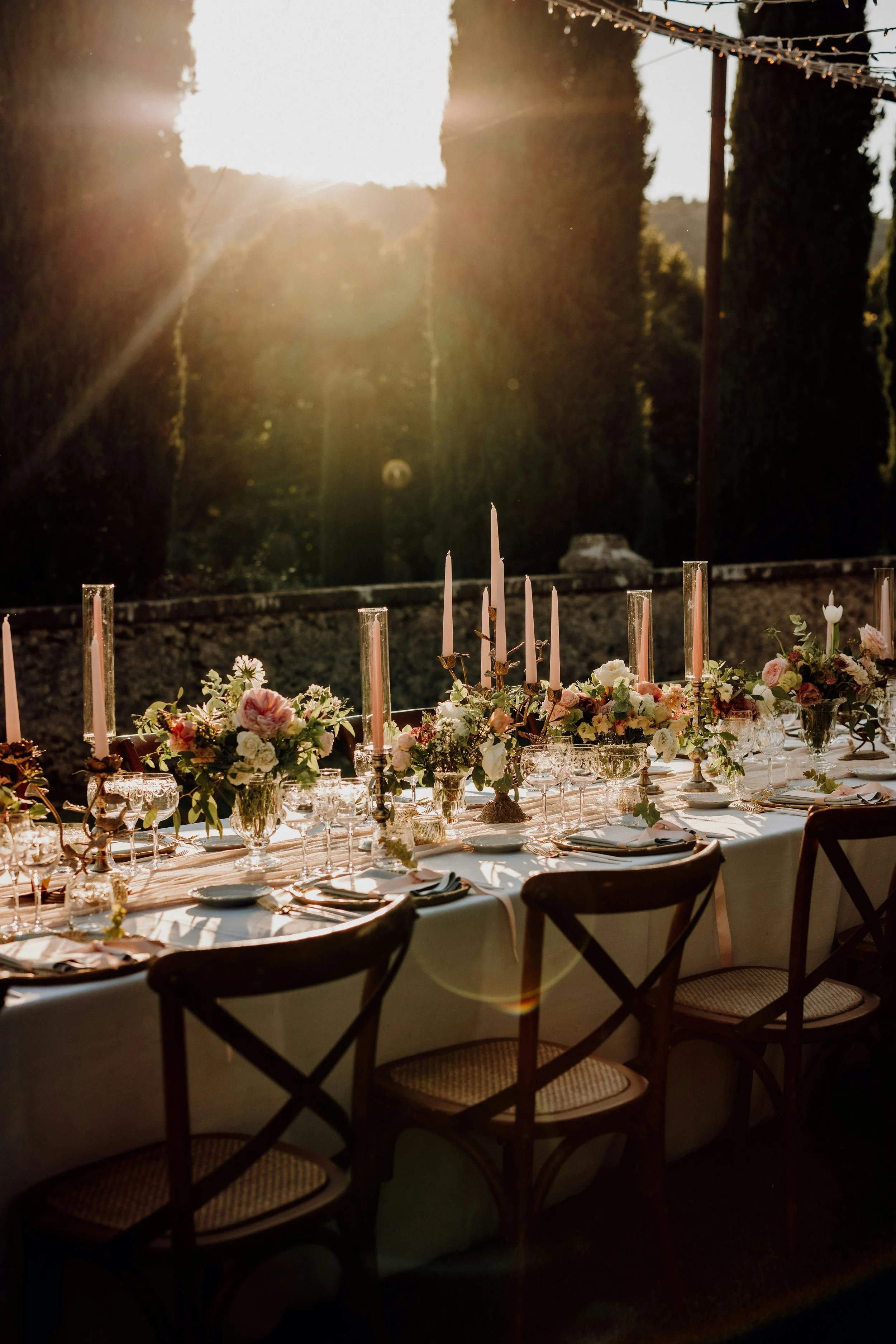 An outdoor dining table decorated with flowers and candles, set in a garden with tall trees and string lights, during sunset.