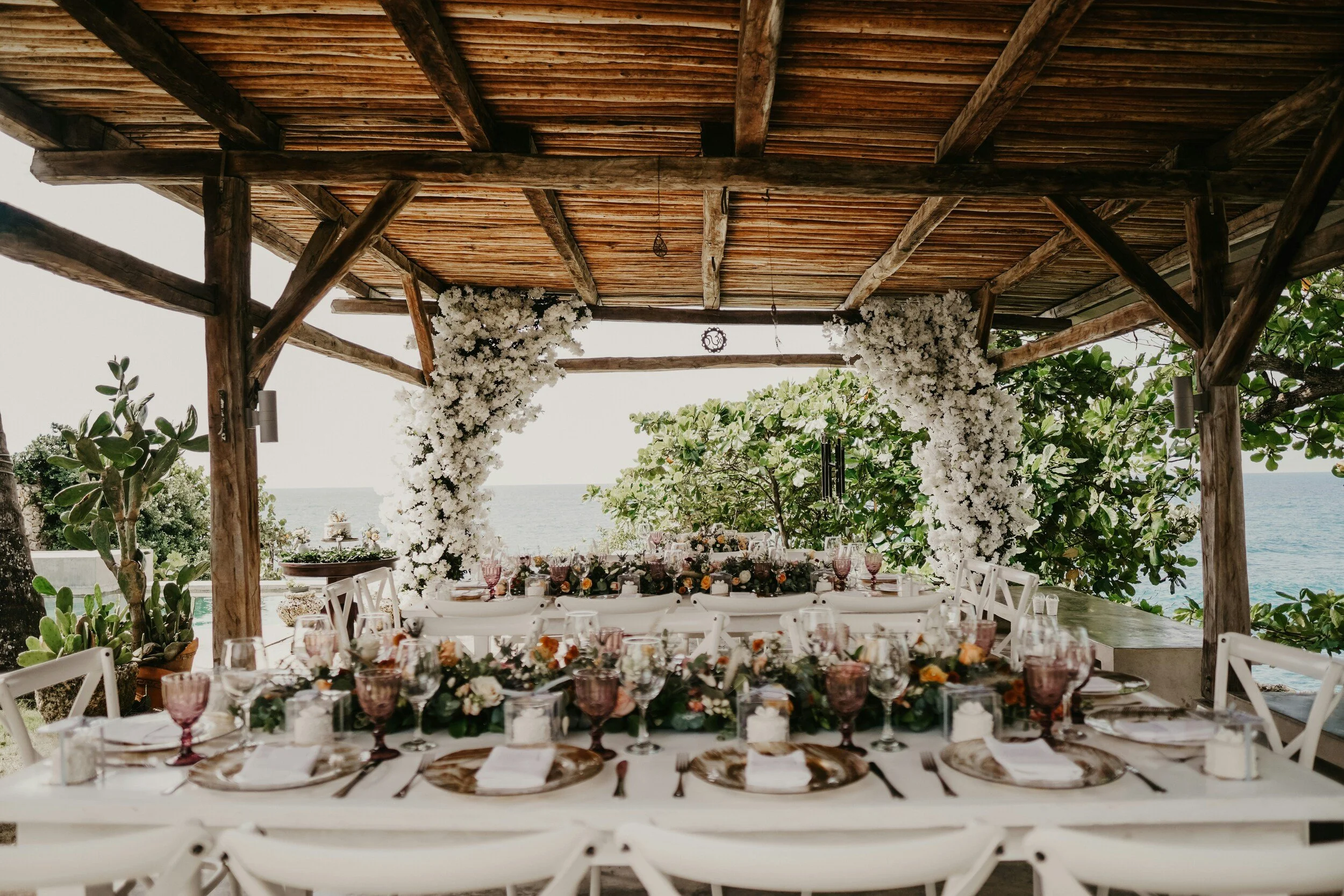 A beautifully decorated outdoor dining area with a long table set with plates, glassware, and flowers, under a wooden pergola overlooking the ocean, with lush greenery surrounding.