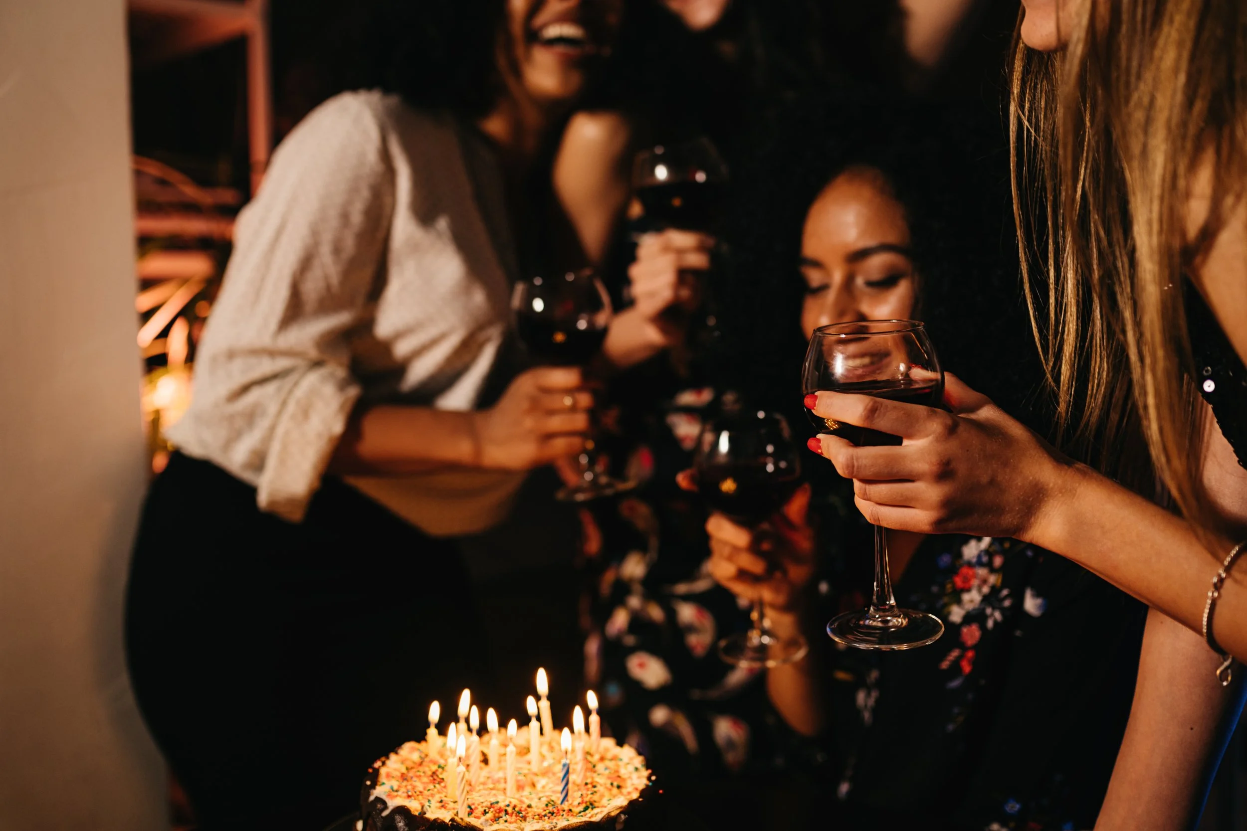 Group of women celebrating a birthday with cake and wine, smiling and toasting in a dimly lit room.