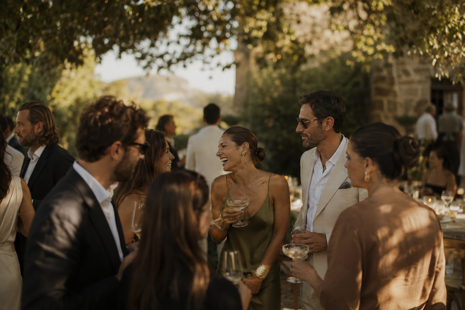 People at an outdoor gathering, dressed in formal attire, socializing and enjoying drinks in a lush garden setting.