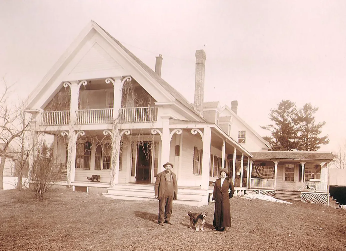 An old black-and-white photograph of a large house with Victorian architectural details, featuring a man and woman standing on the front lawn with a dog, and leafless trees in the background.