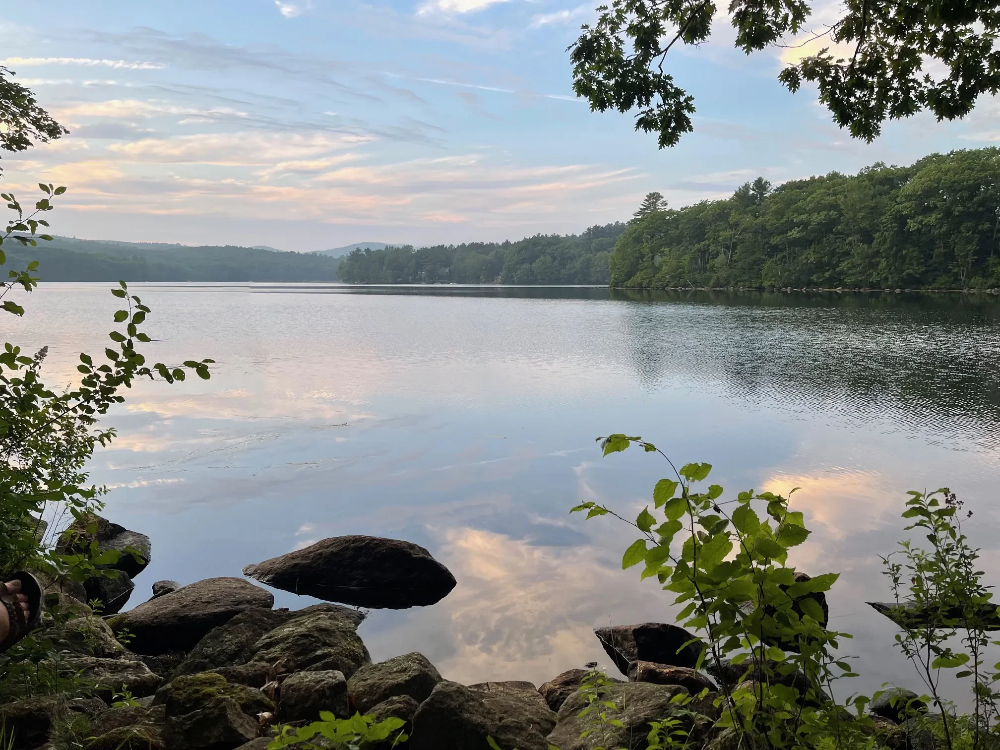 A calm lake surrounded by green trees with a cloudy sky reflected in the water, rocks and green foliage in the foreground.