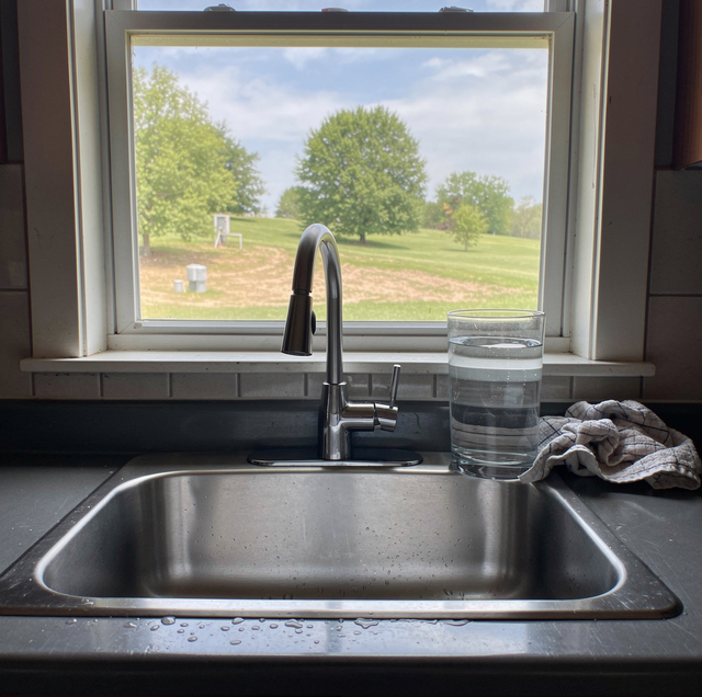 Kitchen countertop with sink, faucet, glass of water, bottle, and cloth towel near a window overlooking a house.