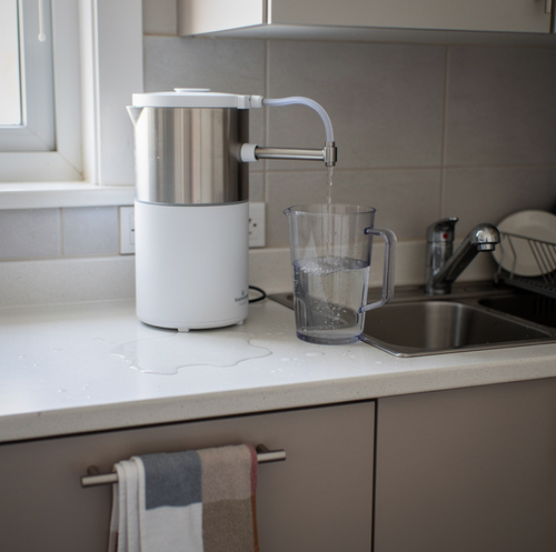 A stainless steel container and glass pitchers sit on a dark kitchen counter next to folded towels near a window.