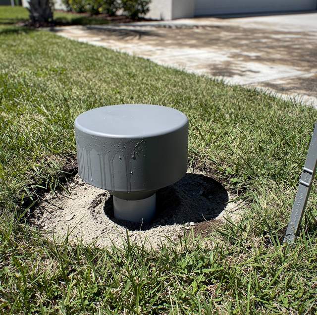 A gray cylindrical cap sits in grass and soil near a driveway with pavement and shrubs visible.