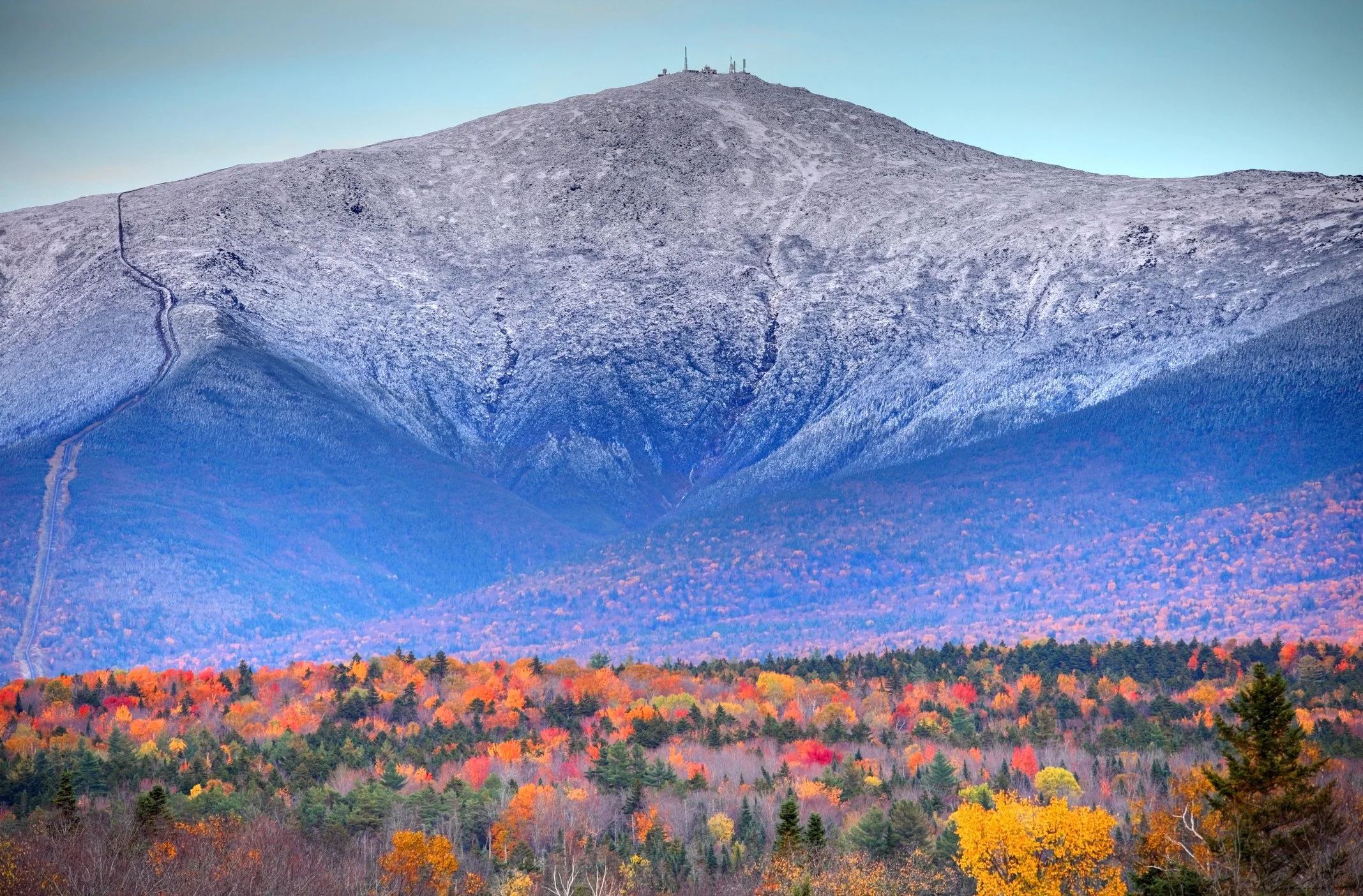A mountain with a road winding up its side, topped with communication towers, surrounded by colorful autumn trees.
