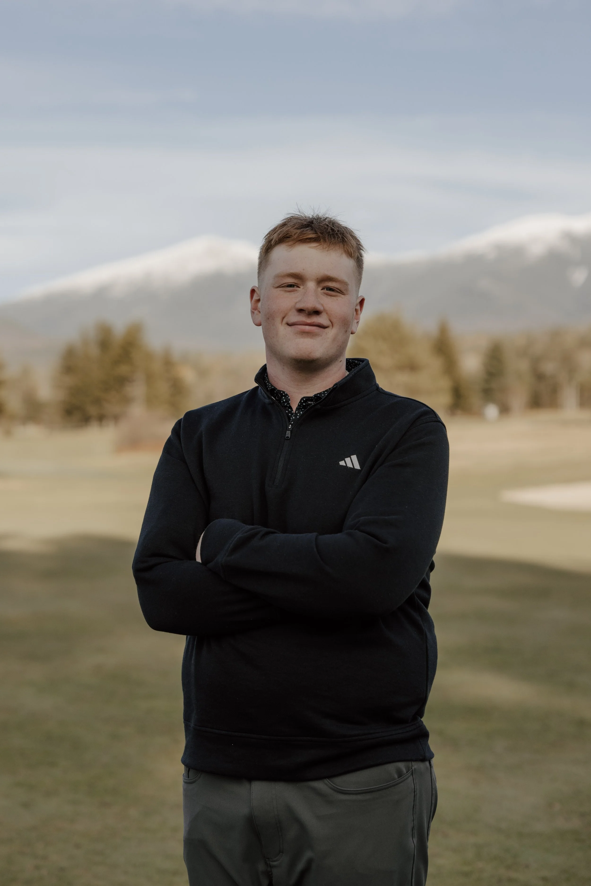 A young man with short reddish hair standing outdoors on a grassy field with trees and snow-capped mountains in the background. He is wearing a black zip-up jacket and gray pants, with arms crossed and a slight smile.