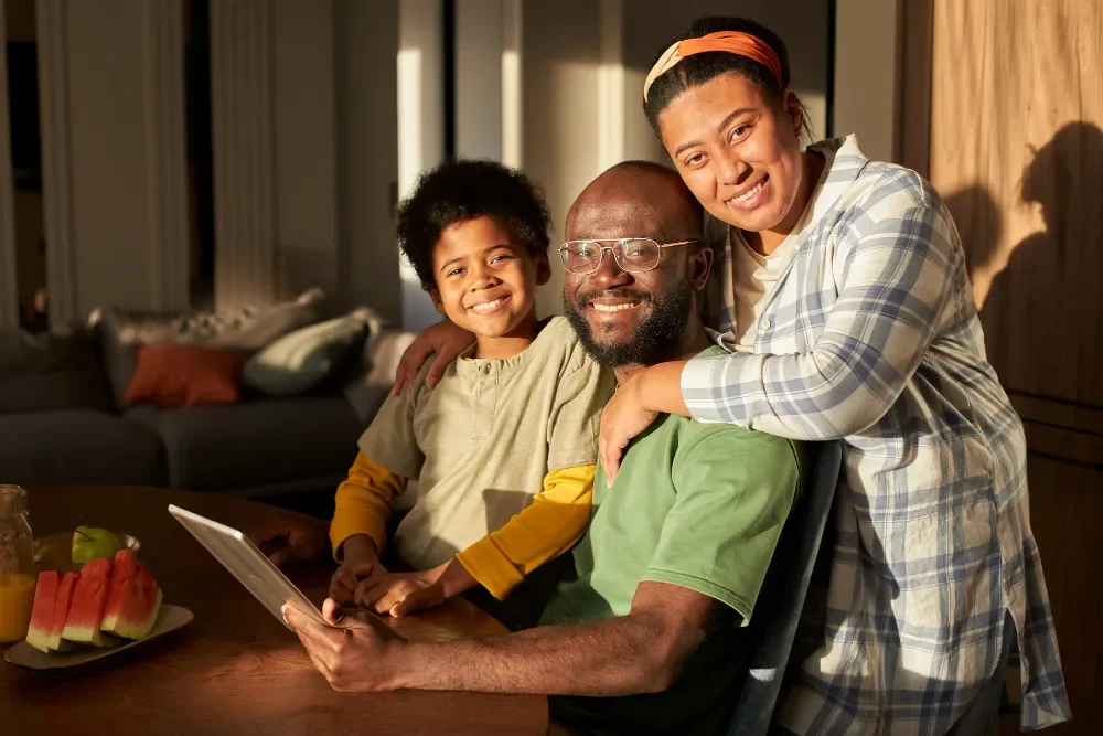 A happy African American family of three, with a father, mother, and young son, sitting together at a dining table in a cozy home, smiling at the camera.