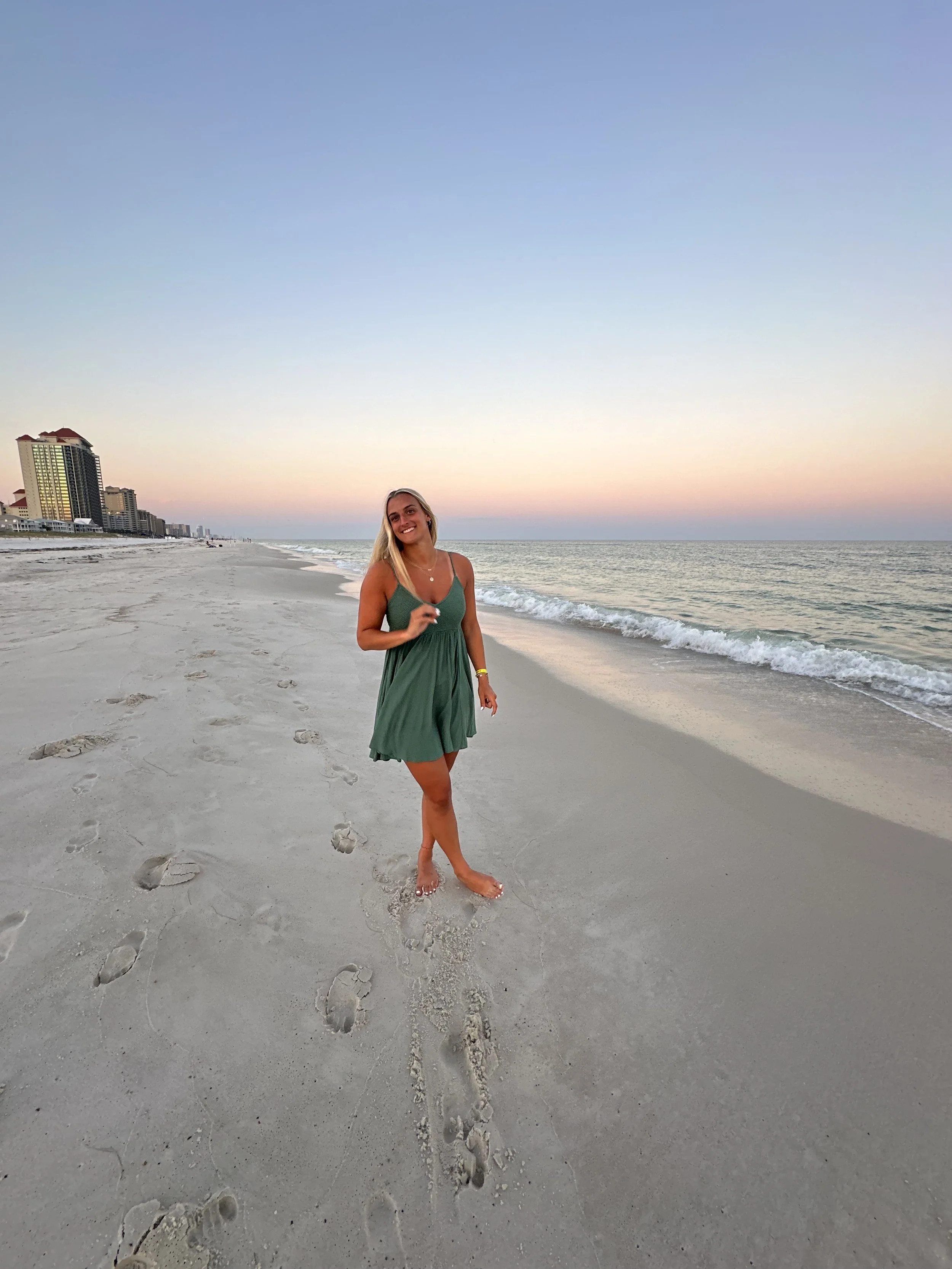 A woman with blonde hair wearing a green dress walking barefoot on a sandy beach near the ocean during sunset, with high-rise buildings in the background.