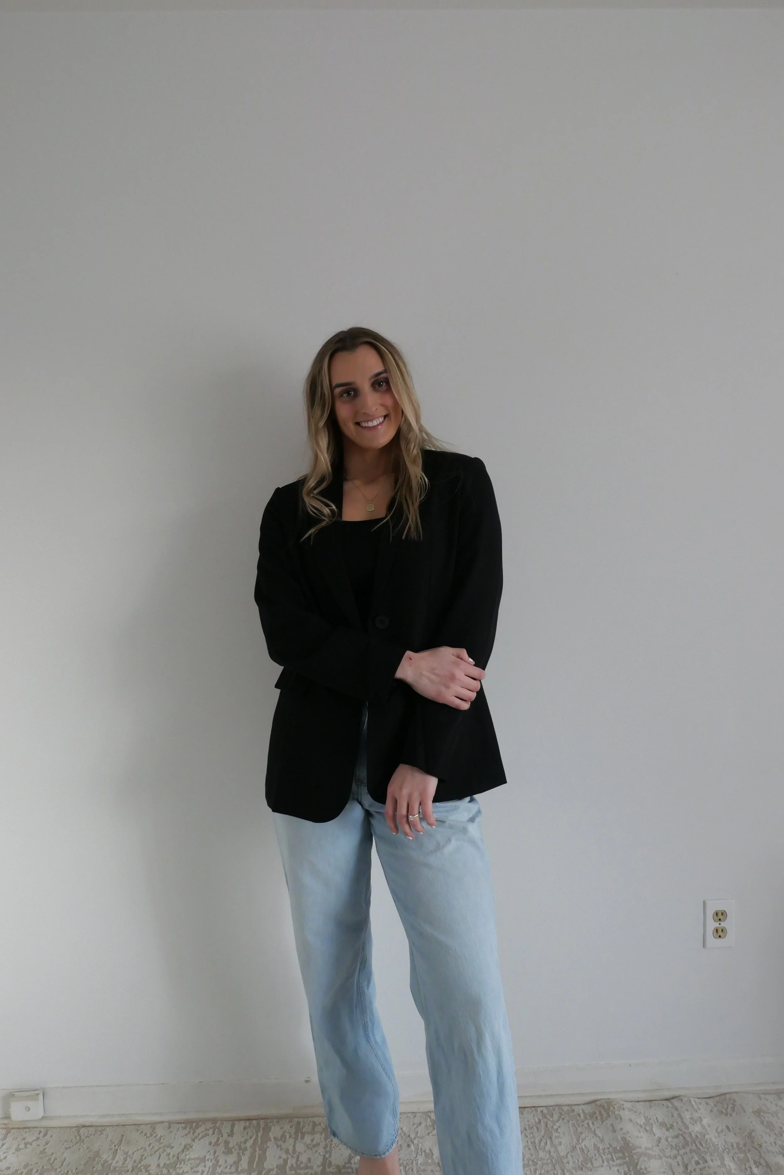 A woman with blonde hair wearing a black blazer, black top, and light blue jeans, standing against a plain beige wall with electrical outlets visible near the floor.