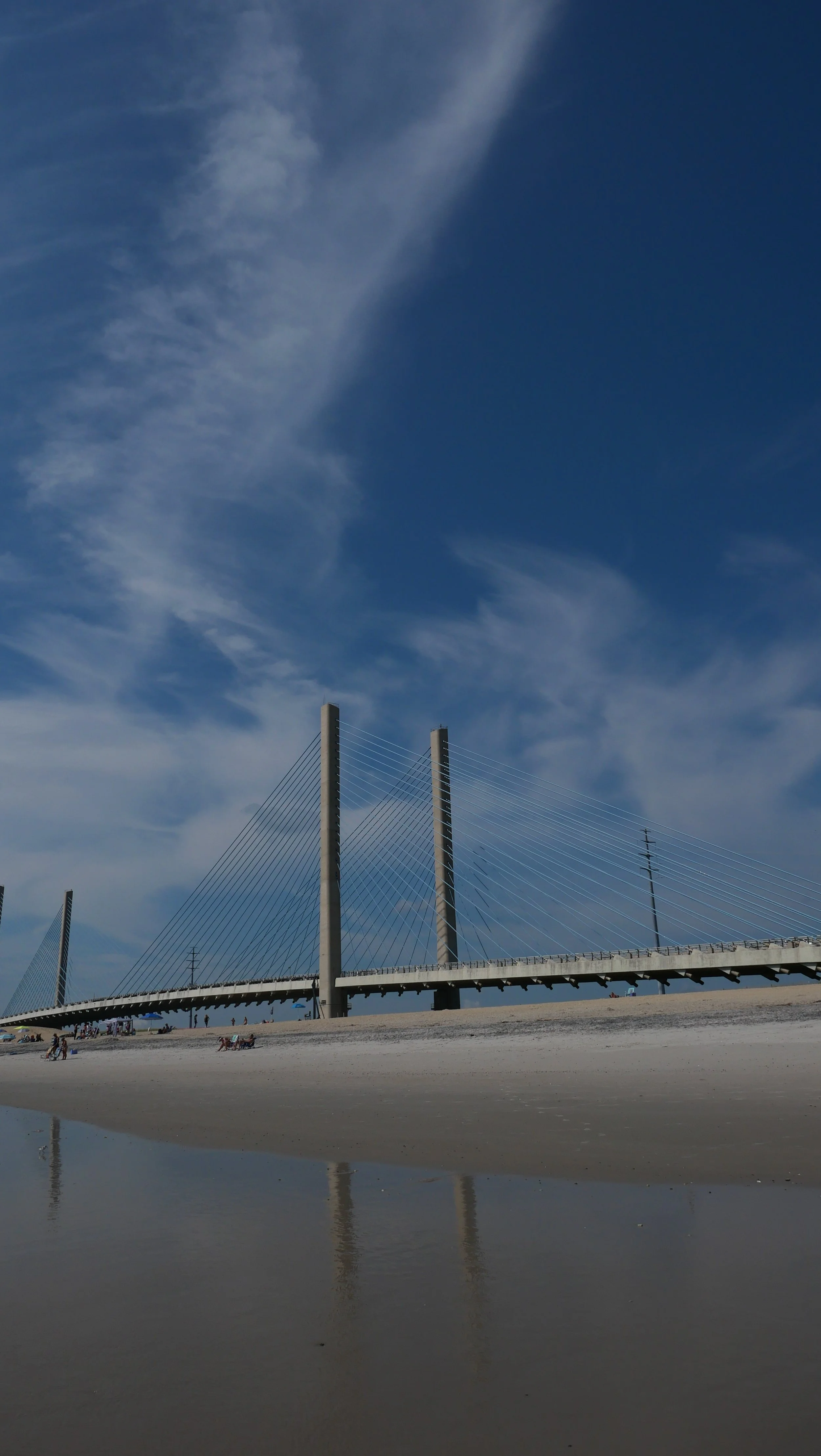 Bridge with tall cable-support towers over a sandy beach, reflected in a shallow water pool. The sky is blue with wispy clouds.