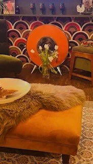 A bouquet of yellow tulips in front of a large framed photograph of a woman with dark hair, on a colorful wall with a patterned background. The scene includes furniture such as a fuzzy blanket on an orange ottoman, a green armchair, and a wooden side