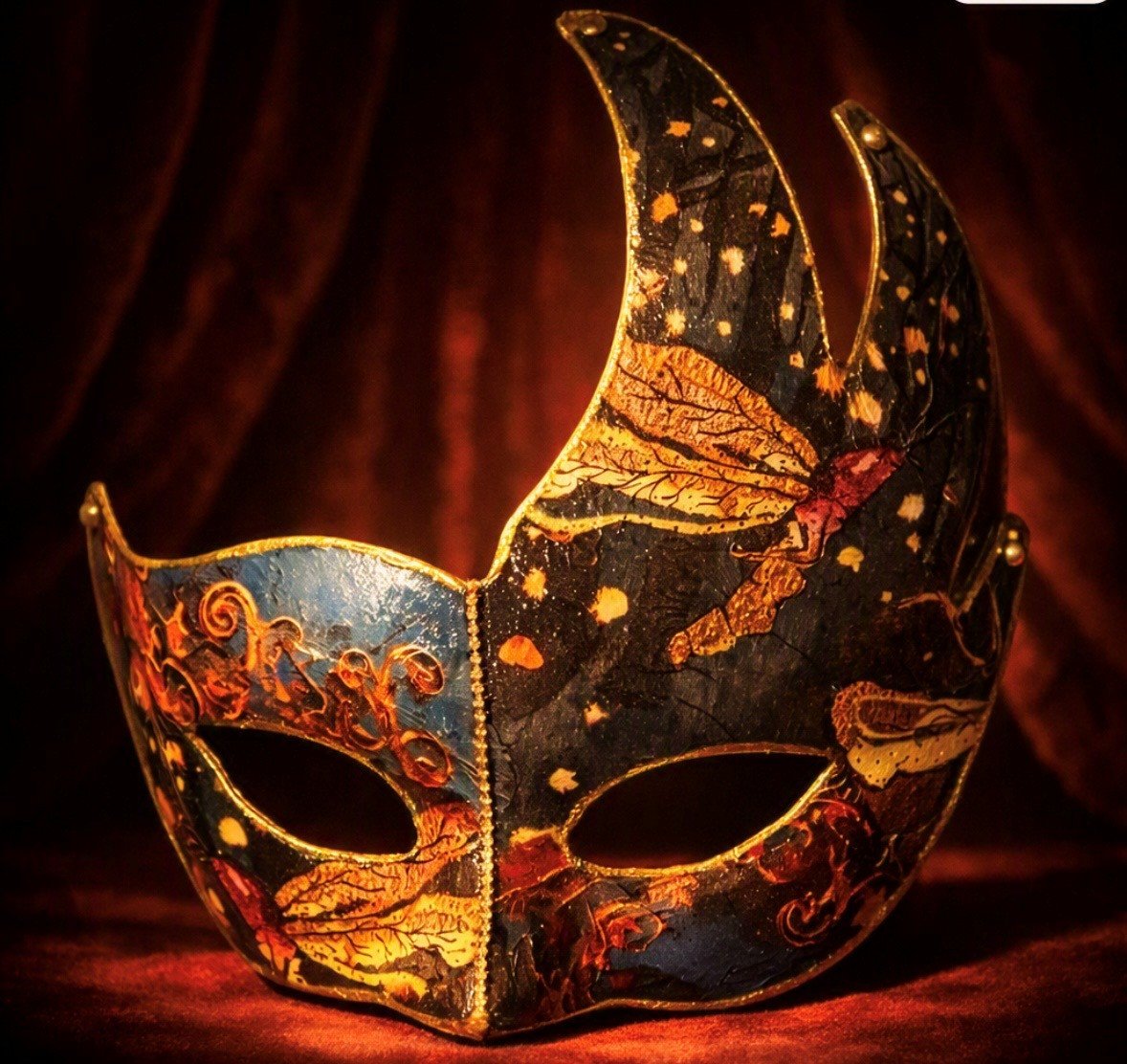 A decorative jester mask with a black and gold leaf design, illuminated with warm lighting against a dark background.