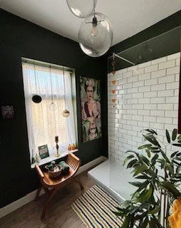 Cozy corner of a room with a large potted plant, a wooden bench, a striped rug, a white brick wall, and a window with sheer curtains, decorated with colorful hanging ornaments.