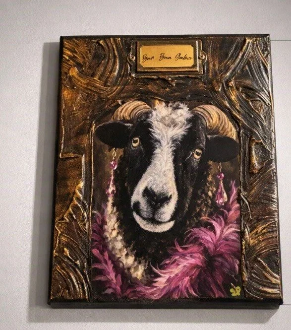 Painting of a black and white sheep with pink earrings and a pink feathered collar, framed with a decorative dark brown frame, with a small gold plaque at the top that says 'Dear Boa Jaber'.
