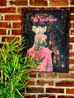 Street art of a zebra wearing sunglasses and a pink jacket, with pink text above that reads 'No ego shit', on a brick wall, partially obscured by green plants in the foreground.