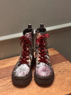 Pair of combat boots with floral embroidery and red laces, placed on wooden floor against a gray wall.