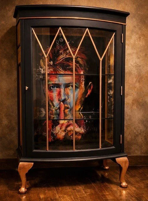 A black wooden display cabinet with a glass door, featuring an abstract colorful portrait of a man with finger to lips painted on the inside back panel.