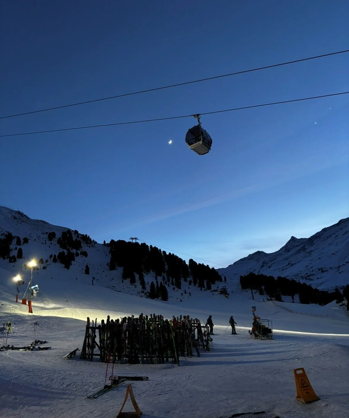 Ski resort at dusk with a gondola lift in the sky, snow-covered mountains, and skiers on the ground, illuminated by artificial lights.
