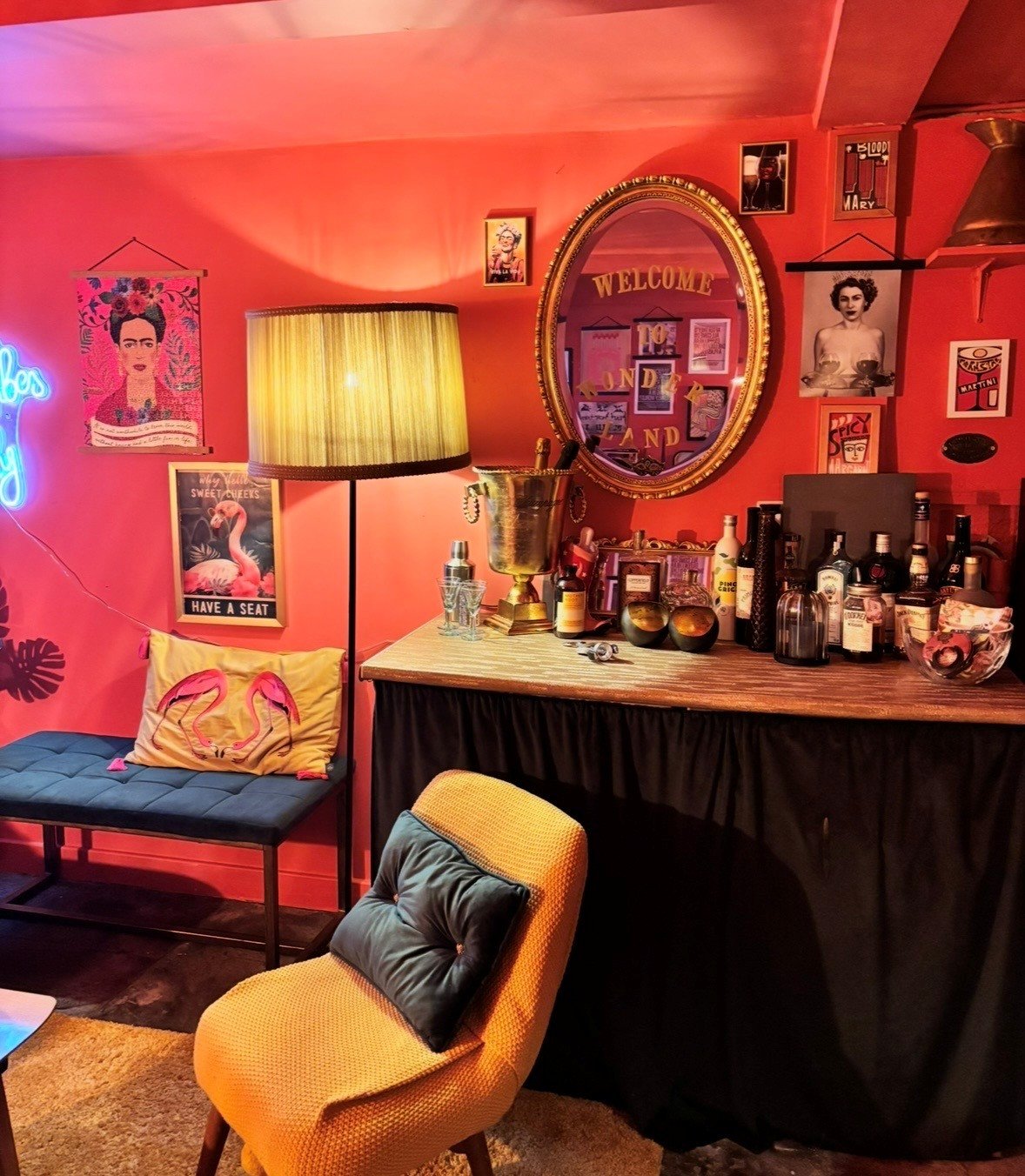 Cozy bar area with pink walls decorated with framed artwork and photographs, a large round mirror, a lamp with a yellow shade, and various bottles of liquor on the wooden bar counter. A yellow armchair with a black cushion and a bench with a flamingo