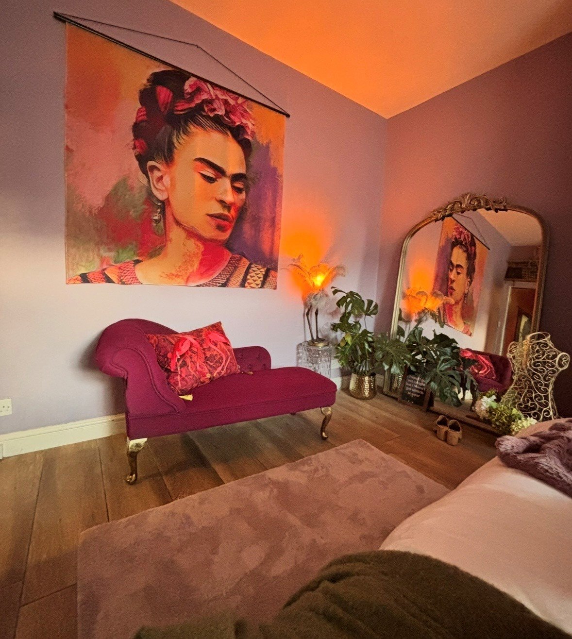Living room with a large Frida Kahlo portrait hanging on a lavender wall, a pink Victorian-style loveseat with decorative pillows, a large mirror reflecting part of the room, potted plants, and warm lighting creating a cozy atmosphere.
