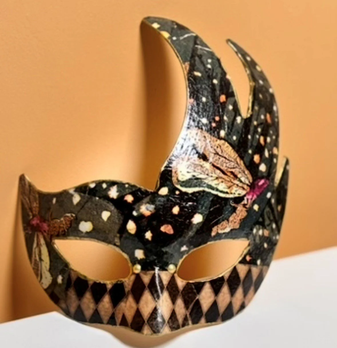 A decorative masquerade mask with a celestial and butterfly design, featuring black, gold, pink, and beige colors, against a peach-colored background.