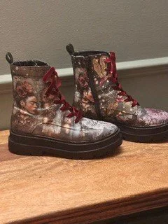 A pair of leather boots with artistic painting or print, featuring a woman, placed on a wooden surface against a gray wall.