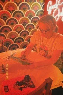 A woman with glasses reading a book or magazine at a table in a retro-themed cafe with colorful, patterned wall decor and illuminated sign.