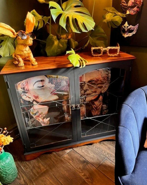 A black display cabinet with glass doors showcasing two large posters of a woman and an elderly man. The cabinet is topped with large green monstera leaves, a small ceramic rabbit figurine, and a pair of glasses. Part of a blue upholstered chair is v