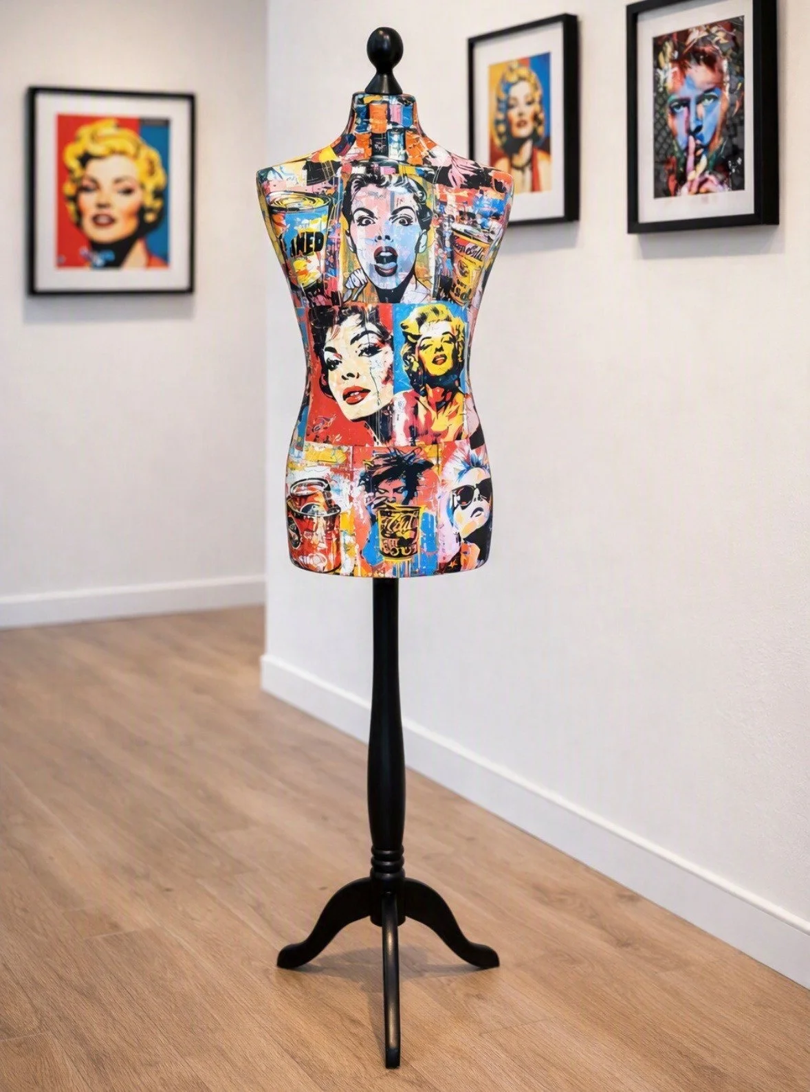Colorful mannequin torso with pop art prints of women, placed on a black stand in an art gallery. Framed pop art portraits hang on the white wall behind.