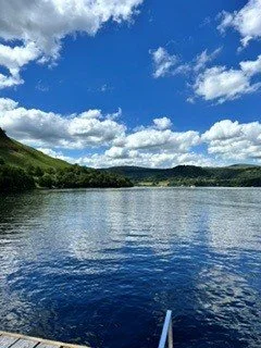 A large body of water with surrounding green hills and a partly cloudy blue sky.