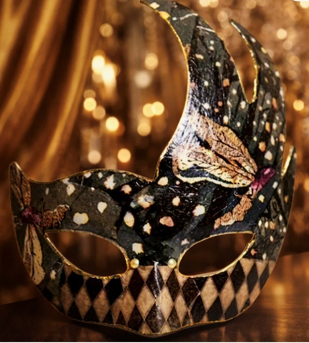 A decorative masquerade mask with a black, gold, and beige pattern, featuring floral and diamond designs, set against a warm, glowing background.